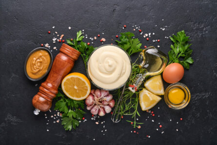 Homemade mayonnaise sauce and ingredients lemon, eggs, olive oil, spices and herbs, black background copy space. Food cooking background. Top view.